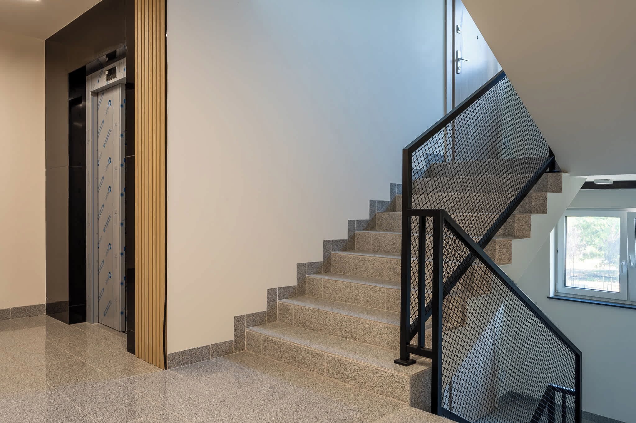 Una escalera moderna con barandilla de metal negro y vistas al exterior a través de un ventanal.