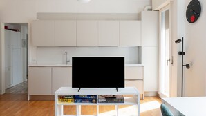 Una cuina moderna amb armaris blancs, un televisor negre sobre un suport blanc i una estant amb jocs de taula.