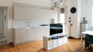 Una cuina moderna amb armaris blancs, televisió integrada i una petita estant amb objectes.