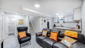 A modern living room with a black leather sofa and armchair, a kitchen with white cabinets and a stainless steel refrigerator, and a staircase leading to another level. - Large family home near Hidden Lake (Denver)