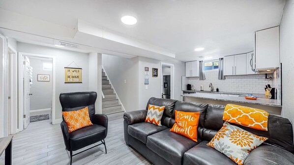 Una sala moderna con un sofá y un sillón de cuero negro, una cocina con armarios blancos y un refrigerador de acero inoxidable, y una escalera que sube a otro nivel.