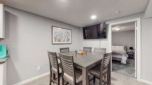 A dining area with a round table, six chairs, a TV mounted on the wall, and a bed in the adjacent room.