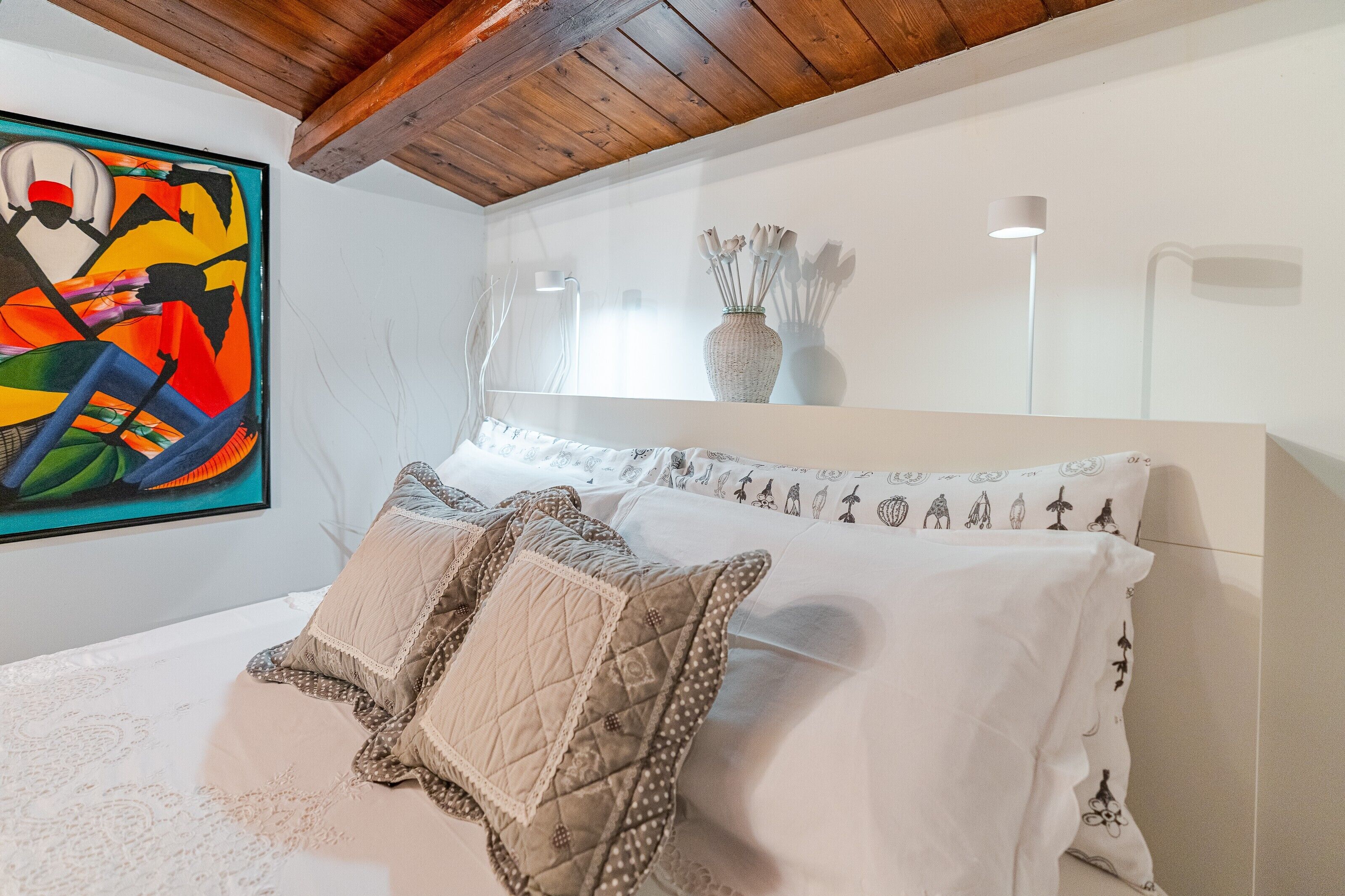 A bedroom with a bed, pillows, a painting, a vase, and a lamp.
