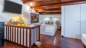 Quarto com teto de madeira, cama com roupa de cama branca, televisão fixada na parede e uma pequena mesa com objetos decorativos.