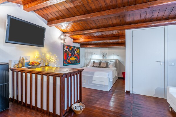 Una habitación con techo de madera, una cama con ropa blanca, un televisor montado en la pared y una mesita con adornos.