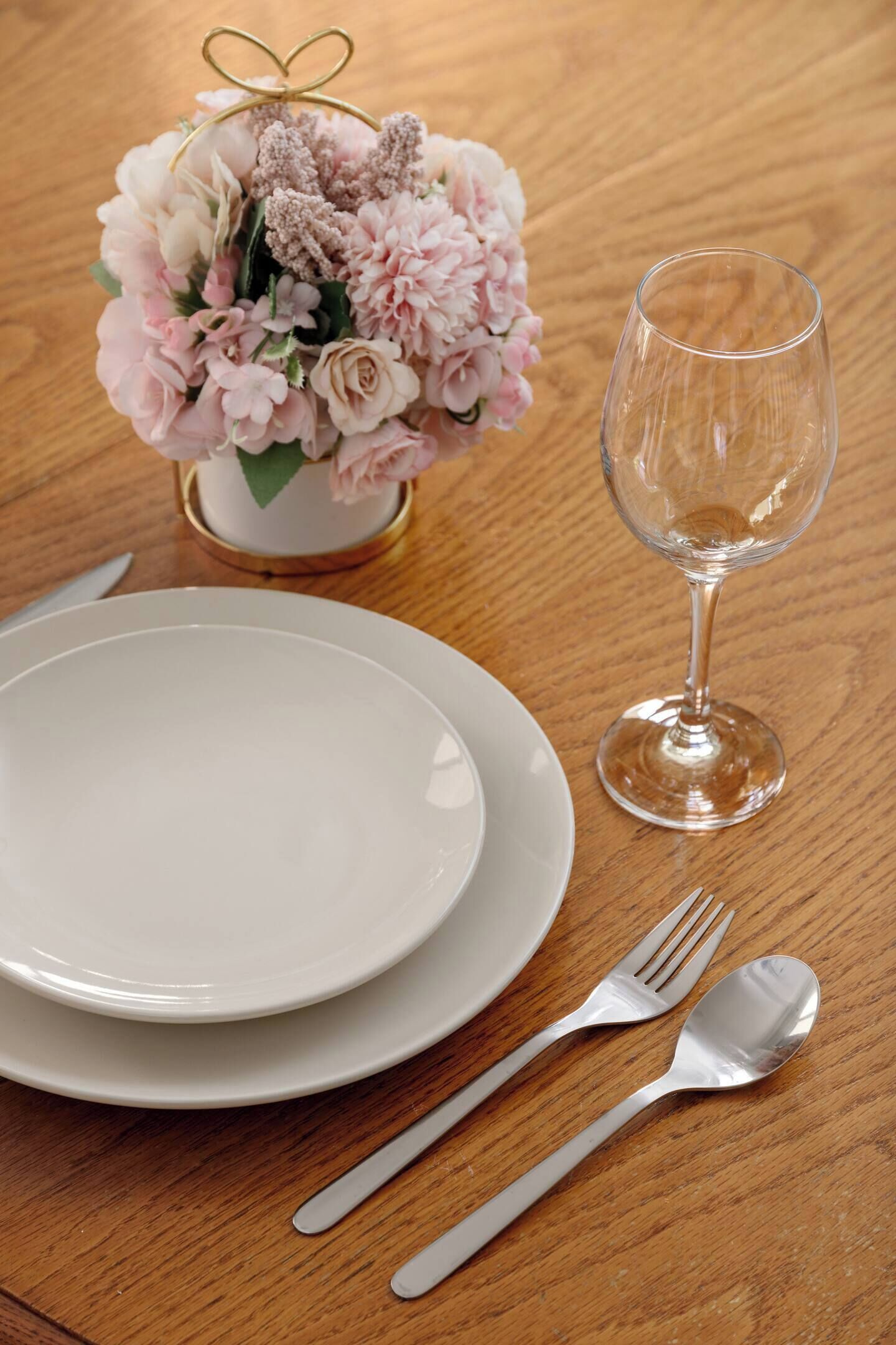 Une table est dressée avec de l’assiette blanche, un verre à vin transparent, une fourchette et une cuillère. Un bouquet de fleurs roses est placé dans un petit vase.