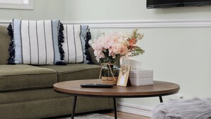 Ein Wohnzimmer mit einem grünen Sofa, einem runden Couchtisch, einem flauschigen Hocker und einem an der Wand montierten Flachbildfernseher.