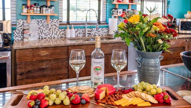 Una encimera de cocina con una botella de vino, copas, una bandeja de frutas y un jarrón con flores.