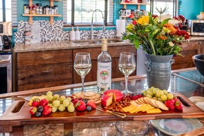 Una encimera de cocina con una botella de vino, copas, una bandeja de frutas y un jarrón con flores.