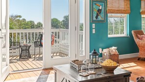 Una habitación con terraza de madera, una puerta blanca que da a la terraza, una pared color verde azulado, una silla de mimbre, una mesa con un cuenco de palomitas de maíz y un cuadro con la imagen de un pájaro.
