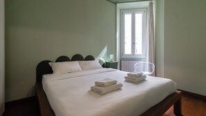 Ein ordentlich bezogenes Bett mit gefalteten Handtüchern, ein Nachttisch mit Lampe und ein Fenster mit Vorhängen.