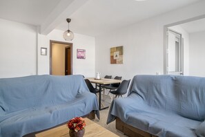 A room with two blue sofas, a wooden coffee table with a vase, a dining table with chairs, and a painting on the wall. - Valdelagrana Beach & Pool Apartment with Shared Pool, Private Terrace, and Wi-Fi (El Puerto de Santa María)