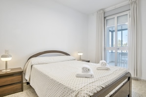 A neatly made bed with a white bedspread, two bedside lamps, and a window with curtains. - Valdelagrana Beach & Pool Apartment with Shared Pool, Private Terrace, and Wi-Fi (El Puerto de Santa María)