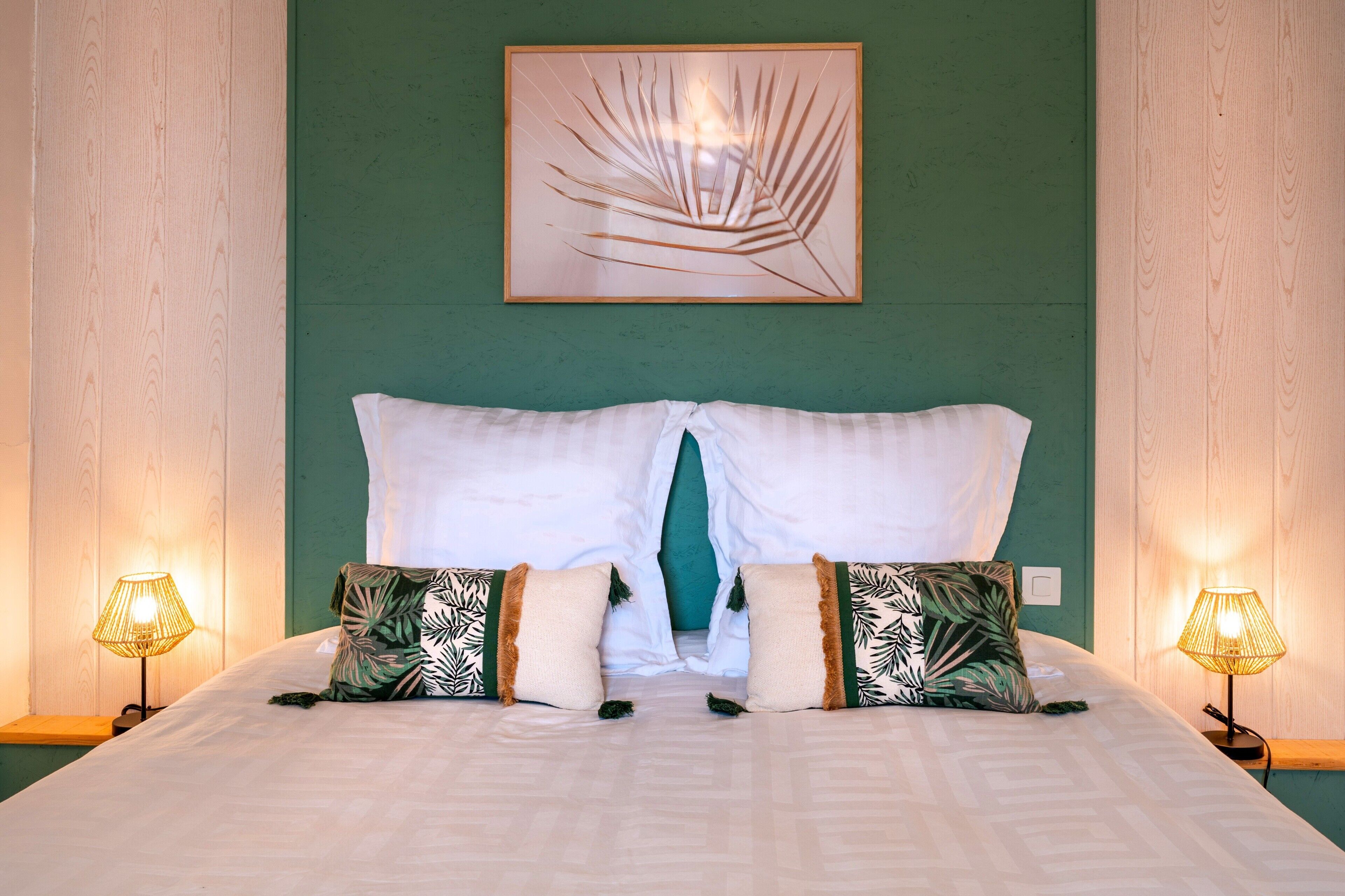 Una cama bien hecha con sábanas blancas, un cojín de acento color turquesa y dos cojines decorativos con diseño de hojas.