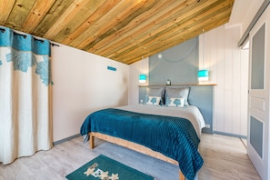 A bedroom with a wooden ceiling, a bed with a teal bedspread, a green rug, and two wall-mounted lights.