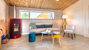 Un salon moderne avec un plafond en bois, un poêle en bois rouge, un canapé agrémenté de coussins à motifs, une lampe et un fauteuil.