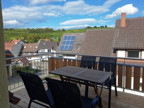 Un balcón con mesa y sillas, un panel solar y vistas a viñedos y casas.
