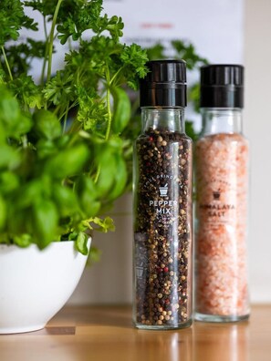 Two glass bottles with labels, one labeled 'PEPPER MIX' and the other 'HIMALAYA SALT', placed on a wooden surface with a white bowl of green herbs in the background. - Trolltunga Hostel (Ullensvang)