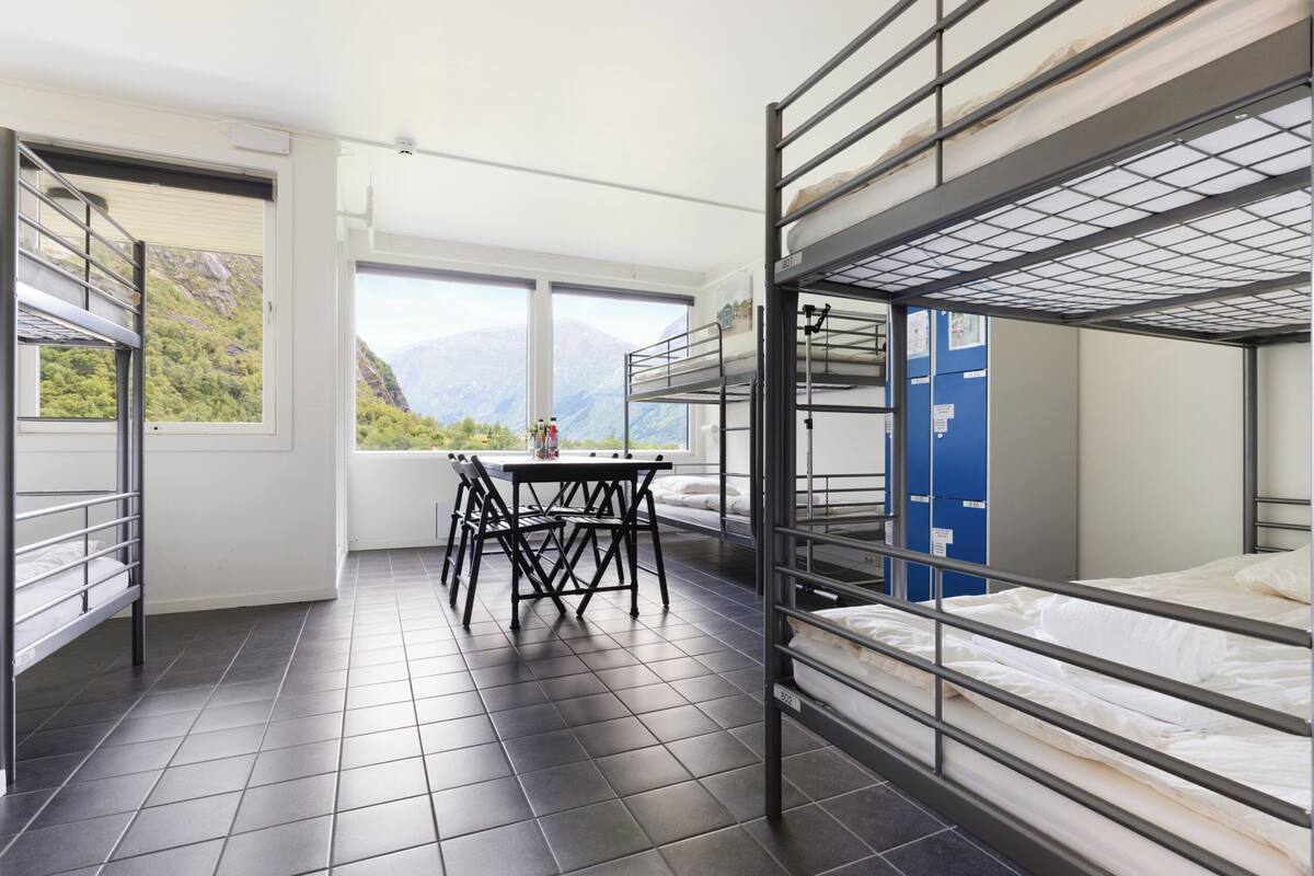 A dormitory room with bunk beds, a table, chairs, and a window with a mountain view.