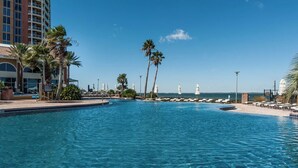 Ein klares, blaues Schwimmbecken mit Liegestühlen und Sonnenschirmen, umgeben von Palmen und einem Gebäude mit einem Balkon.
