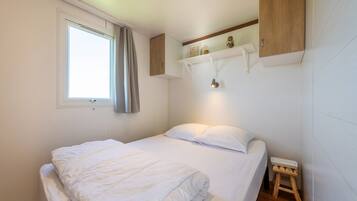 En säng för en person med vita sängkläder, en liten träpall, ett fönster med gardiner och en hylla med dekorativa föremål.