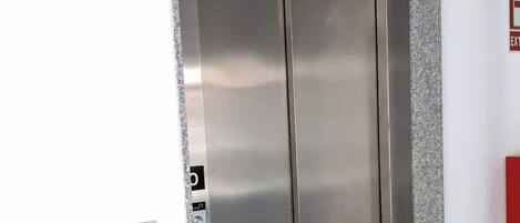 Un ascensor con acabado metálico, un panel de control con botones y un detector de incendios en la pared.