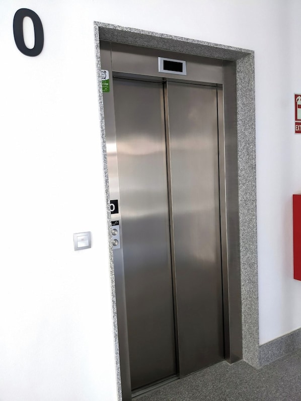 An elevator with a metallic finish, a control panel with buttons, and a fire alarm on the wall.