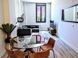 A modern living room with a glass dining table, brown chairs, a black sofa, and a large window.