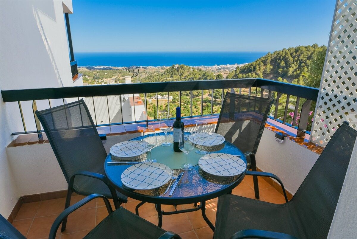 A balcony with a table set for four, overlooking a coastal landscape.