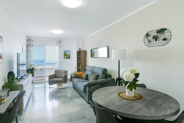 A modern living room with a grey sofa, a dining table with chairs, and a view of the sea through large windows.
