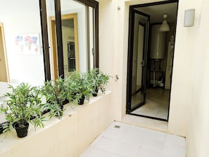 A narrow corridor with potted plants lining the wall, leading to a room with a visible water heater and a hanging light fixture.