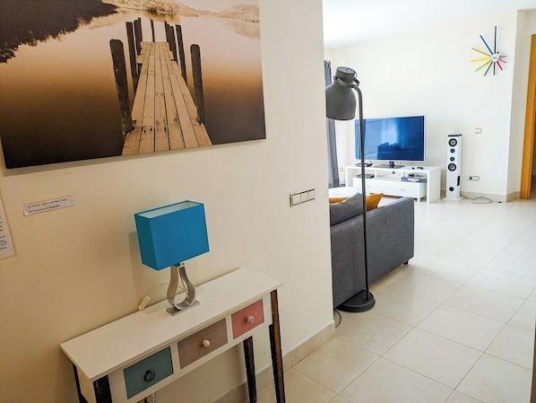 A modern living room with a flat-screen TV, a blue desk lamp, and a large painting of a wooden dock.