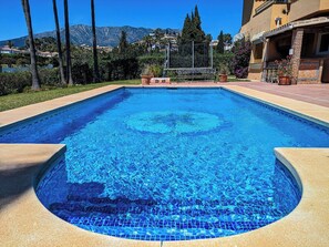A swimming pool with a clear blue water surface, surrounded by a paved deck and a building with a balcony.