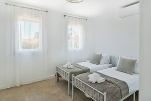 A room with two beds, each with a white mattress and grey bedding, and a window with sheer curtains.