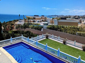 Une piscine entourée d’une clôture blanche, avec vue sur la mer et des bâtiments en arrière-plan.