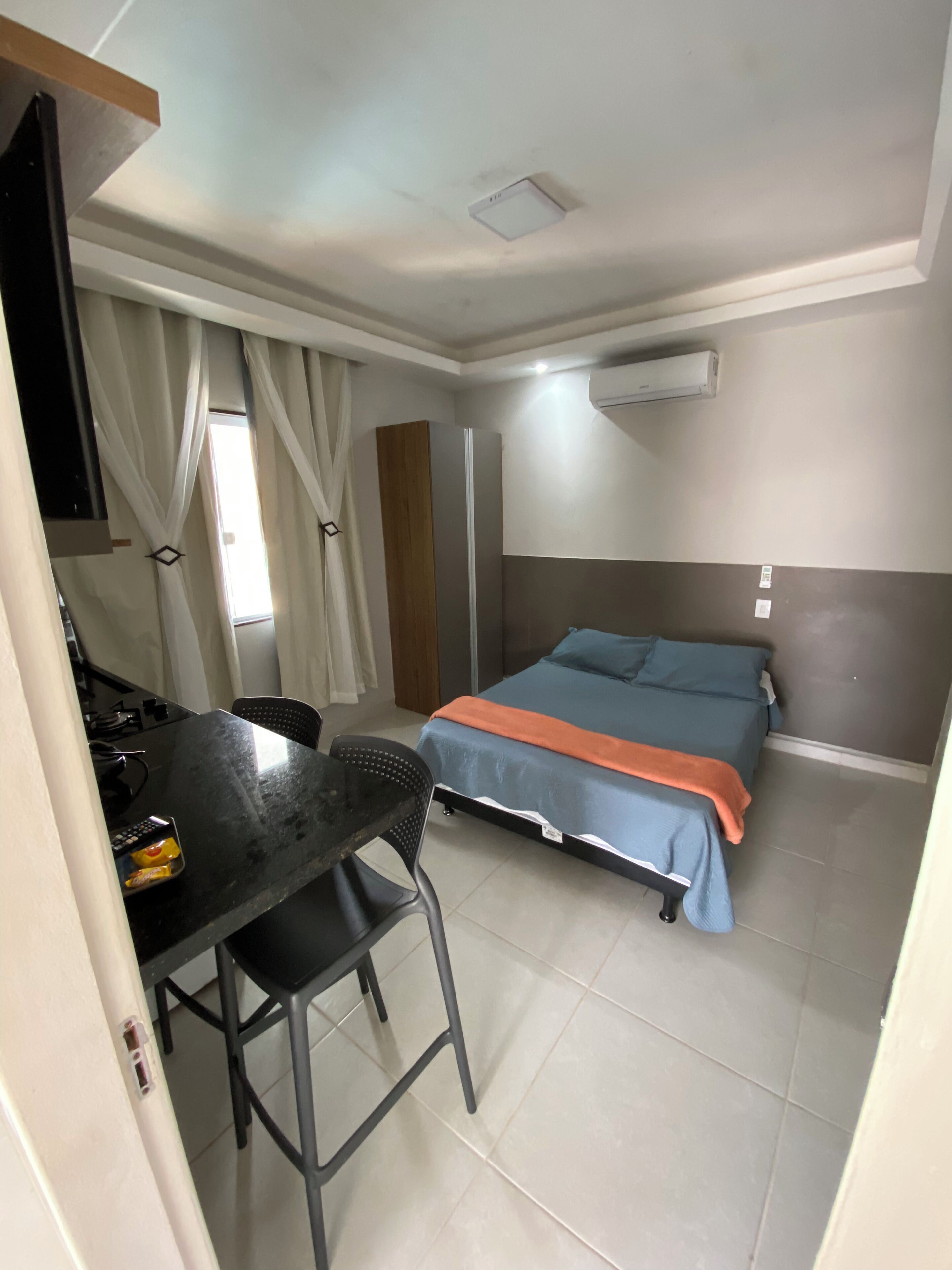 Quarto compacto com uma cama, escrivaninha e cadeira. Há um ar condicionado na parede.