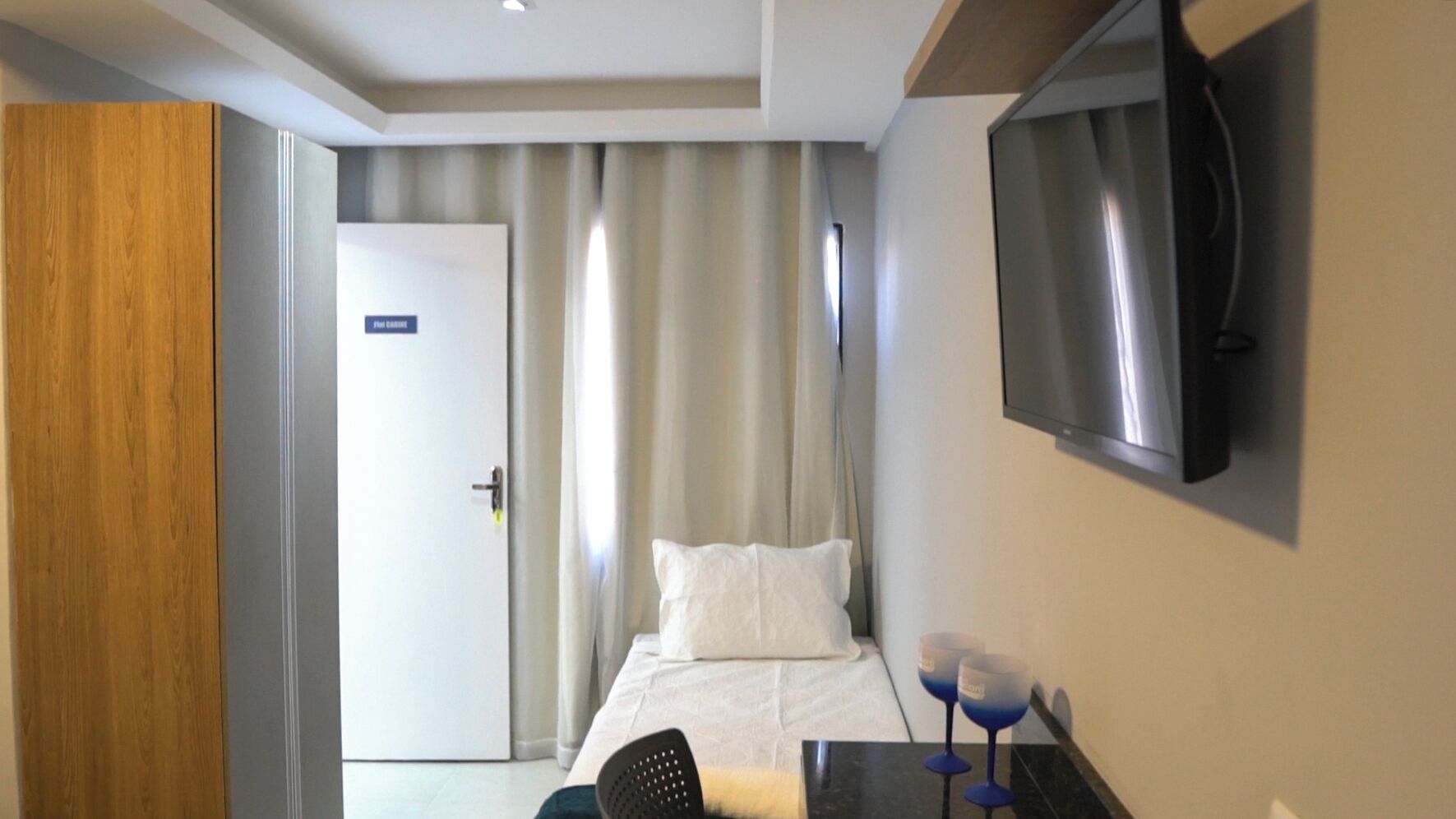 Habitación de hotel con cama, escritorio con dos copas de vino, televisor en la pared y puerta con un cartel.