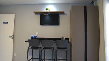 A compact room with a flat-screen TV mounted on the wall, a small shelf with plants, and a large wardrobe. There are three bar stools and a small table with two blue glasses.