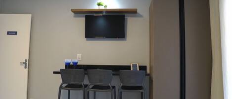 Una habitación compacta con un televisor de pantalla plana montado en la pared, un estante pequeño con plantas y un armario grande. Hay tres taburetes altos y una mesa con dos vasos azules.