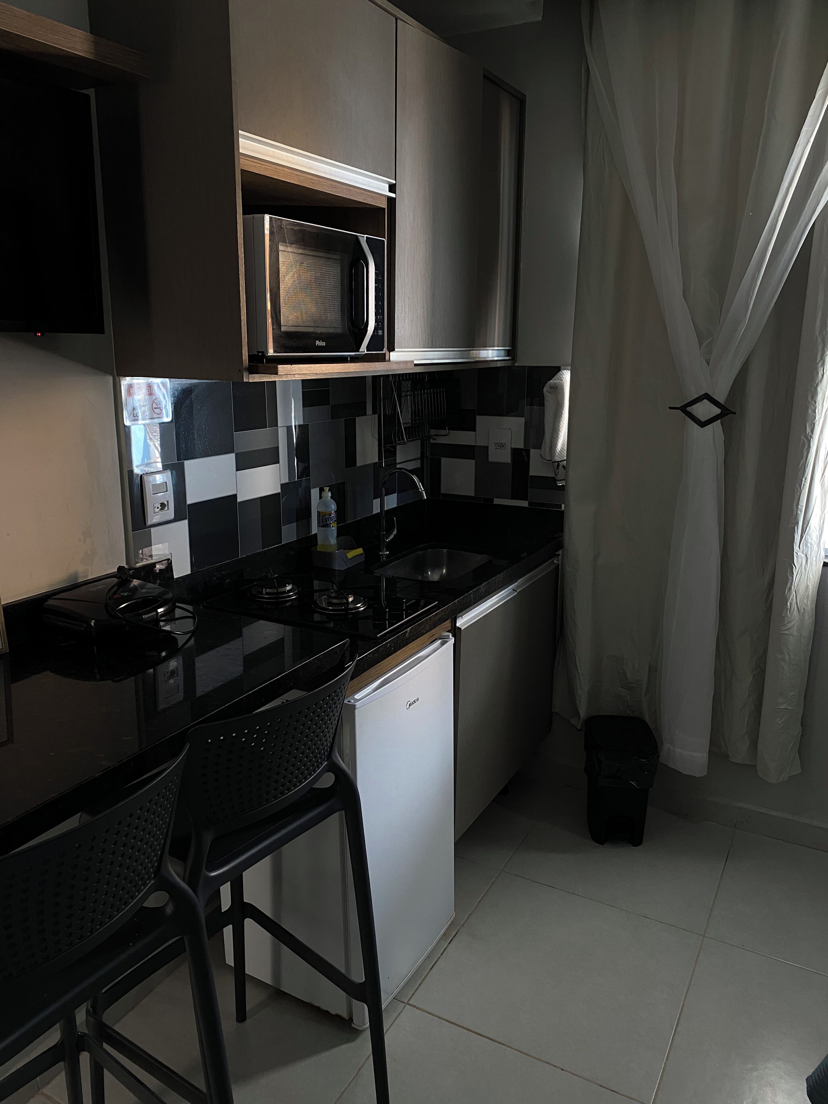 A compact kitchen with a black countertop, a white refrigerator, a microwave, and a sink. There are two black bar stools and a black trash bin.