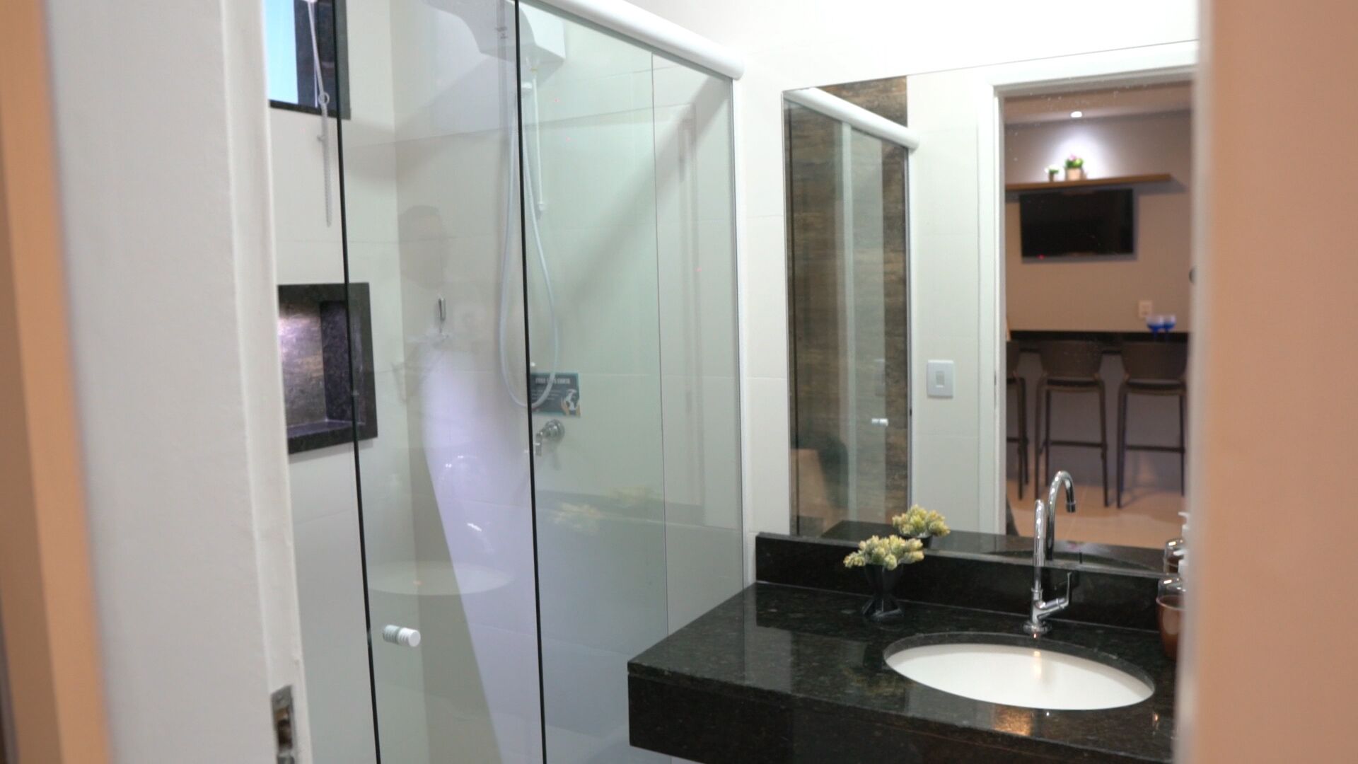 Un baño moderno con cabina de ducha de vidrio, lavamanos de granito negro y un espejo que refleja una zona de cocina.