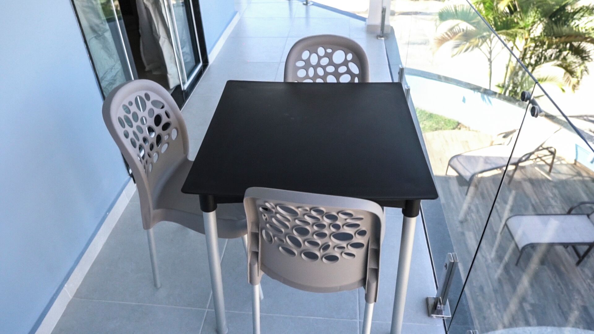 A small outdoor dining area with a table and two chairs, a glass railing, and a view of palm trees.