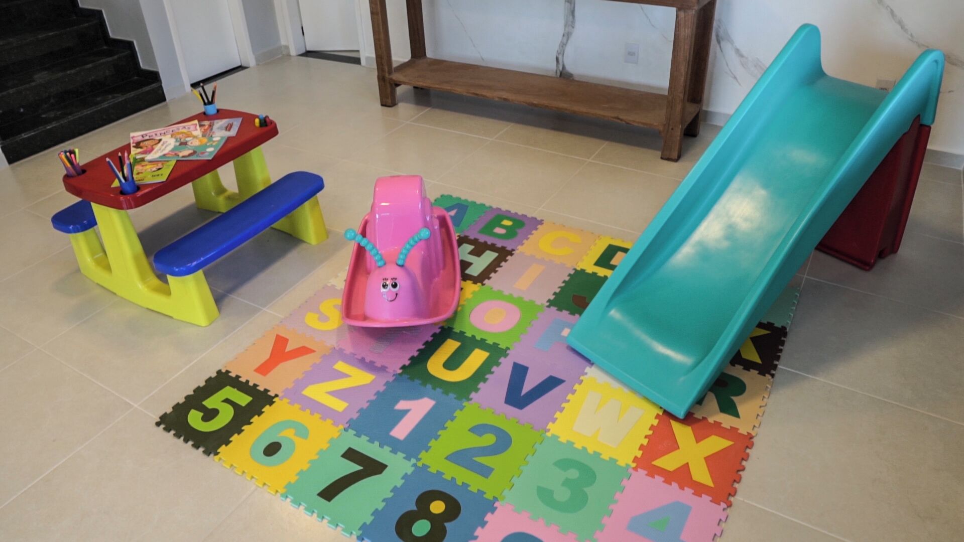 Uma área de recreação com um escorrega, uma mesinha com brinquedos e tapetes de espuma com o alfabeto no chão.