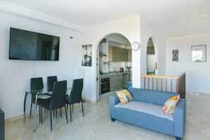 A modern living room with a flat-screen TV, a dining area with chairs, a sofa with colorful cushions, and a kitchen in the background.