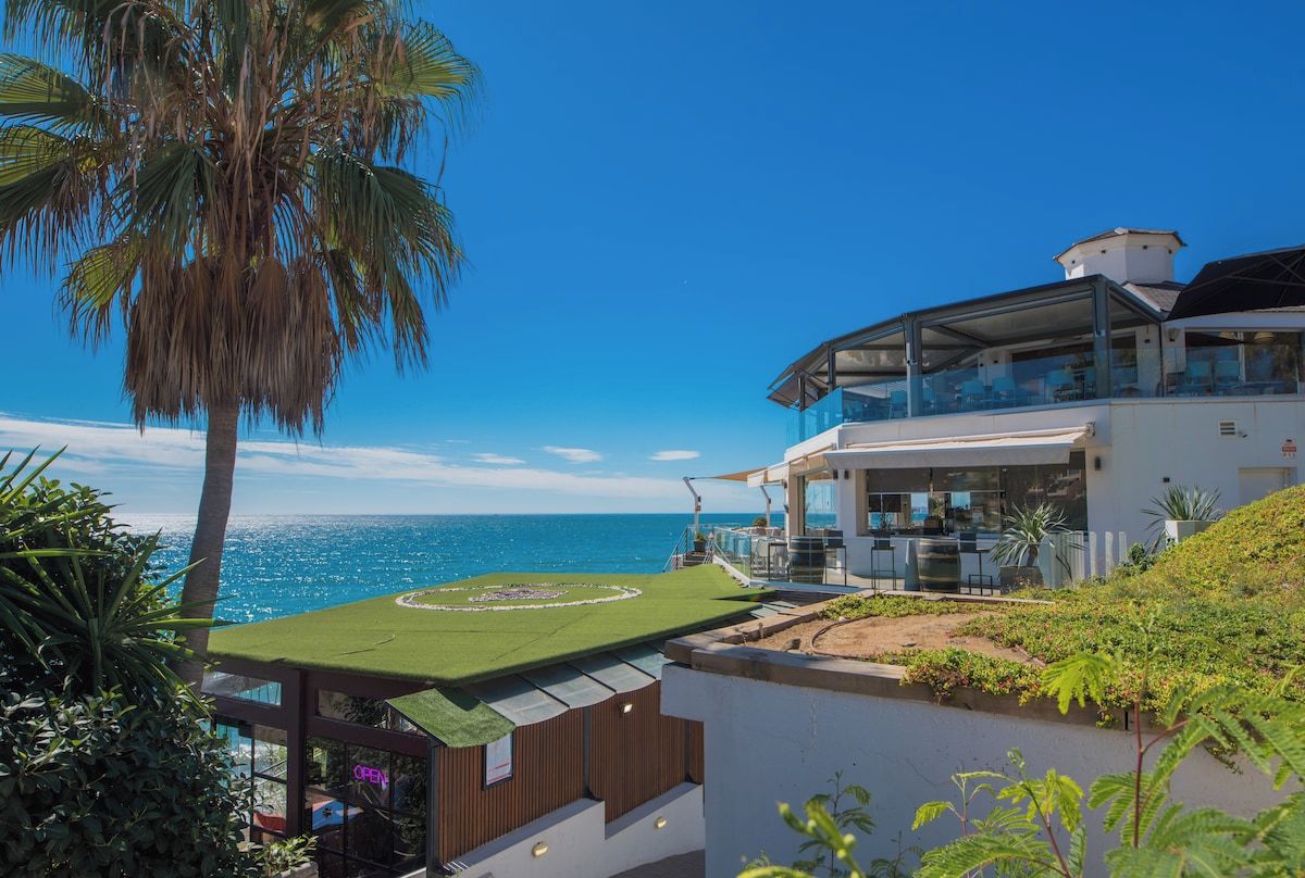 A modern building with a rooftop terrace overlooking the ocean, featuring a circular garden and a glass balustrade.