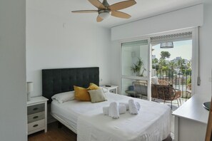 A hotel room with a bed, a nightstand, a desk, a chair, and a balcony with a view of palm trees and buildings.