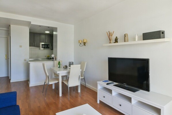 A modern living room with a flat-screen TV, a dining area with a white table and chairs, and a kitchen with grey cabinets and a microwave.