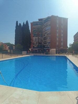 Une piscine avec une terrasse carrelée, entourée d’une clôture, et un immeuble à plusieurs étages en arrière-plan.