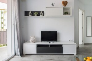 Ett modernt vardagsrum med en platt-TV placerad på en vit TV-bänk, en liten hylla med dekorativa föremål och ett glasbord med frukt.