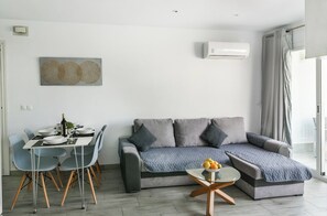 A modern living room with a grey sofa, a dining table with chairs, and a small side table with fruit.